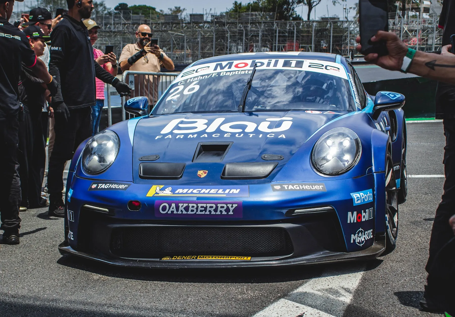 Porsche azul de frente no pit lane, cercado por pessoas da equipe