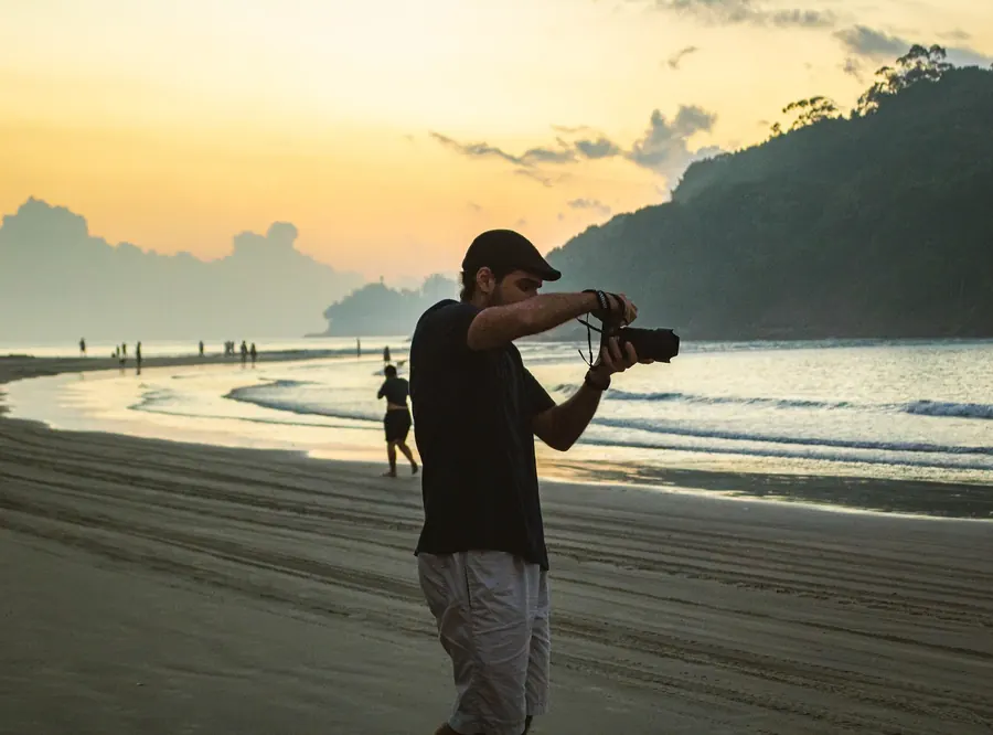 Fotógrafo na areia ao entardecer, com morro ao fundo
