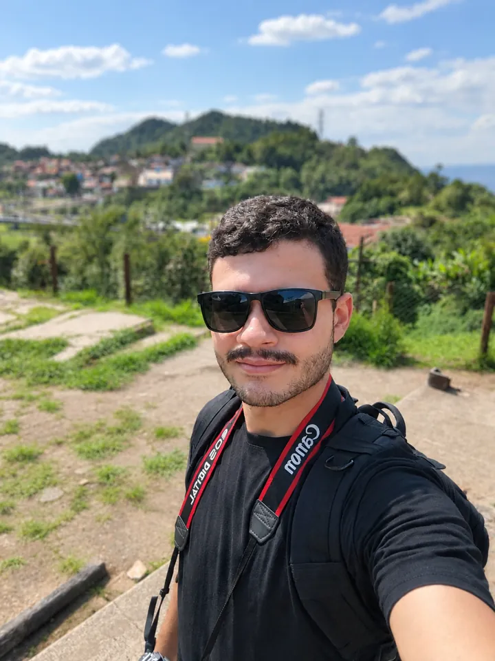 Kauã Nicolas sorrindo, de óculos escuros e camiseta preta, com a alça da câmera no pescoço e um morro ao fundo